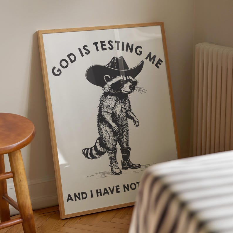 Puede incluir: L&aacute;mina art&iacute;stica enmarcada con un mapache con sombrero y botas de vaquero. La ilustraci&oacute;n en blanco y negro tiene el texto "GOD IS TESTING ME" y "AND I HAVE NOT". La impresi&oacute;n est&aacute; en un marco de madera.