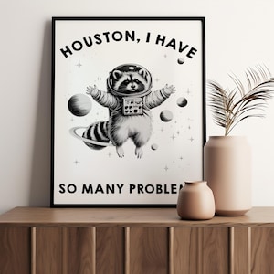 May include: A framed black and white wall art print featuring a raccoon wearing a space suit floating in space. The text reads HOUSTON, I HAVE SO MANY PROBLEMS. The raccoon is surrounded by planets, stars, and a ringed planet in a cosmic scene.