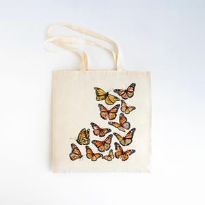 Puede incluir: Un bolso de tela de color crema con un dise&ntilde;o estampado de varias mariposas monarca en tonos naranja, negro y blanco. Las mariposas est&aacute;n dispuestas en un grupo en la parte inferior derecha del bolso, el cual cuenta con dos asas.