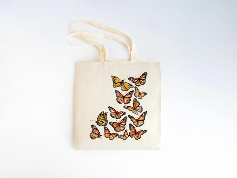 Puede incluir: Un bolso de tela de color crema con un dise&ntilde;o estampado de varias mariposas monarca en tonos naranja, negro y blanco. Las mariposas est&aacute;n dispuestas en un grupo en la parte inferior derecha del bolso, el cual cuenta con dos asas.