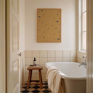 May include: A bathroom scene featuring a white bathtub, a wooden stool, and a framed artwork with small figures on a mustard-colored background. The floor has a black and white checkered pattern, and a towel is draped over the tub.