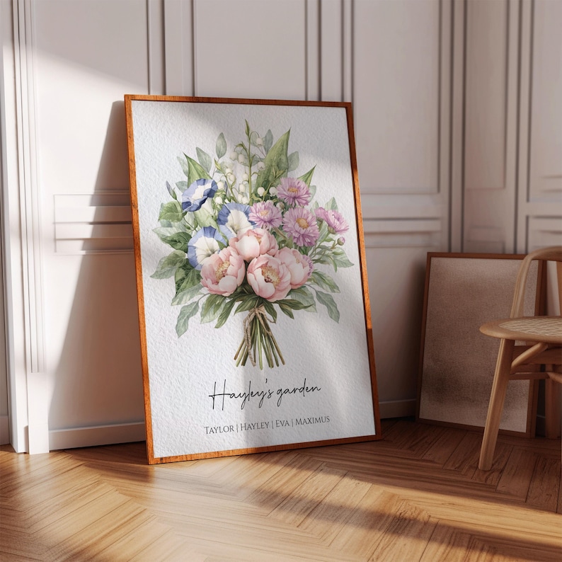 Op de afbeelding: Een ingelijste aquarelprint met een boeket van roze pioenrozen, blauwe winde en paarse dahlia's. De tekst "Hayley's garden" staat onder de bloemen, met namen eronder. De lijst heeft een warme bruine kleur.