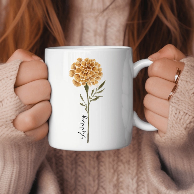 May include: White ceramic mug with a marigold flower illustration and the name "Ashley" written in cursive.