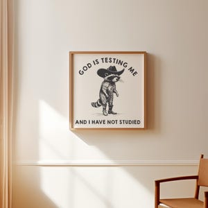 Puede incluir: Un estampado enmarcado con una ilustraci&oacute;n en blanco y negro de un mapache con sombrero de vaquero y botas. El texto encima del mapache dice "GOD IS TESTING ME" y debajo "AND I HAVE NOT STUDIED".