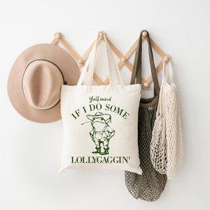 May include: A white tote bag with green text that reads "Y'all mind if I do some lollygaggin'" and a cartoon frog wearing overalls and a hat. The bag is hanging on a wooden rack with a brown hat and two string bags.