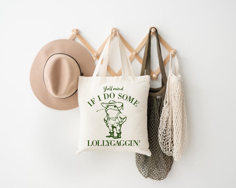 May include: A white tote bag with green text that reads "Y'all mind if I do some lollygaggin'" and a cartoon frog wearing overalls and a hat. The bag is hanging on a wooden rack with a brown hat and two string bags.