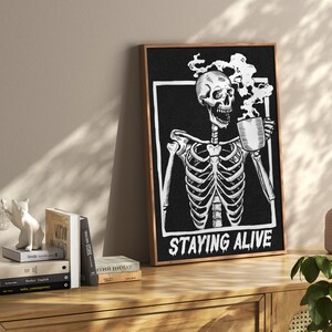 May include: Black and white illustration of a skeleton holding a steaming cup of coffee. The text "STAYING ALIVE" is below the skeleton. The illustration is framed in a dark wood frame.
