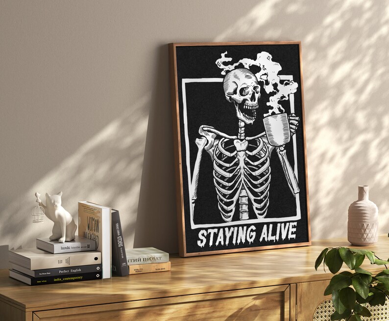 May include: Black and white illustration of a skeleton holding a steaming cup of coffee. The text "STAYING ALIVE" is below the skeleton. The illustration is framed in a dark wood frame.