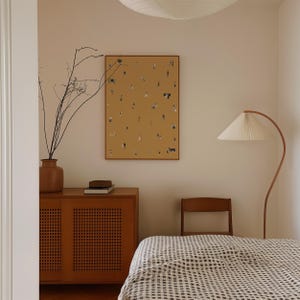 May include: A warm-toned bedroom scene featuring a framed artwork with tiny figures on a mustard background. A wooden cabinet, a vase with branches, and a floor lamp with a pleated shade are visible. The bed has a checkered pattern.