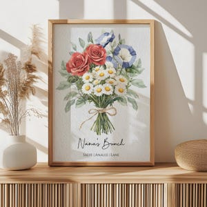 Op de afbeelding: Een ingelijste aquarelprint met een boeket bloemen. Het boeket bevat rode rozen, blauwe en witte winde en witte madeliefjes. De woorden "Nana's Bunch" staan onder de bloemen, met de namen Sadie, Analee en Lane.
