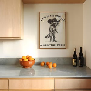Puede incluir: Escena de cocina con un p&oacute;ster enmarcado con un mapache con sombrero de vaquero, con el texto "GOD IS TESTING ME AND I HAVE NOT STUDIED". Un cuenco de terracota lleno de naranjas y dos botellas de vino en la encimera.
