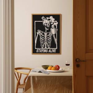 May include: Black and white illustration of a skeleton drinking from a mug with steam rising from the mug. The text "STAYING ALIVE" is below the skeleton.
