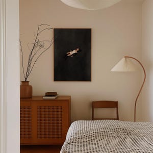 May include: A framed rectangular wall art print featuring a dark charcoal background with a stylized illustration of a figure floating in a white swimsuit. The artwork is displayed on a light beige wall above a wooden cabinet with cane doors.