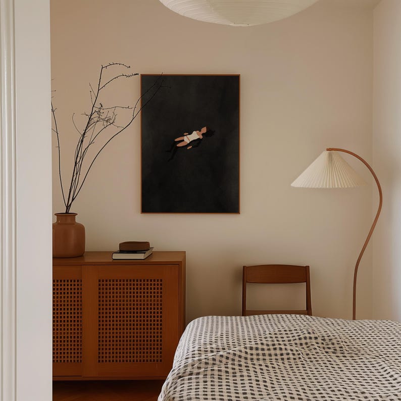 May include: A framed rectangular wall art print featuring a dark charcoal background with a stylized illustration of a figure floating in a white swimsuit. The artwork is displayed on a light beige wall above a wooden cabinet with cane doors.