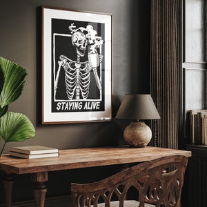 May include: Black and white illustration of a skeleton holding a steaming mug with the text "Staying Alive" printed below. The illustration is framed and hanging on a dark wall.
