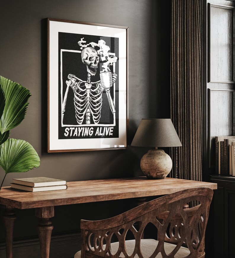 May include: Black and white illustration of a skeleton holding a steaming mug with the text "Staying Alive" printed below. The illustration is framed and hanging on a dark wall.