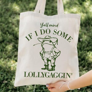 May include: A white canvas tote bag with green text that reads "Y'all mind if I do some lollygaggin'" and a cartoon frog wearing a hat and overalls.