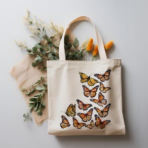 Puede incluir: Un bolso de tela color crema con un dise&ntilde;o estampado de varias mariposas monarca naranjas y negras dispuestas en un grupo en la parte inferior derecha. El bolso tiene dos asas de tela a juego y una forma rectangular sencilla.