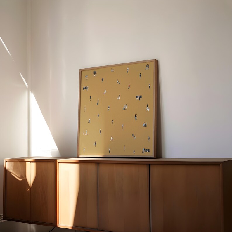 May include: A framed art print featuring a mustard-yellow background with numerous tiny figures scattered across it. The artwork is encased in a light brown wooden frame and rests on a wooden cabinet. The room has a minimalist aesthetic.