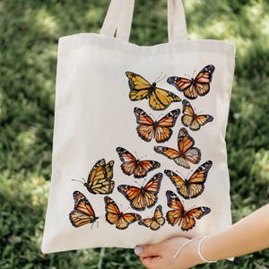 Puede incluir: Una bolsa de tela de color crema con un dise&ntilde;o estampado de varias mariposas monarca en tonos naranja, negro y blanco. Las mariposas est&aacute;n dispuestas en un grupo en la parte frontal de la bolsa, que cuenta con dos asas de tela.