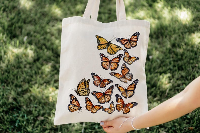 Puede incluir: Una bolsa de tela de color crema con un dise&ntilde;o estampado de varias mariposas monarca en tonos naranja, negro y blanco. Las mariposas est&aacute;n dispuestas en un grupo en la parte frontal de la bolsa, que cuenta con dos asas de tela.