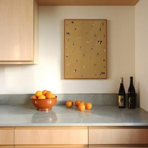 May include: A kitchen scene featuring a terracotta bowl filled with oranges on a gray countertop. A framed artwork with tiny figures hangs on the wall. Two wine bottles stand to the right. Light wood cabinets are visible.