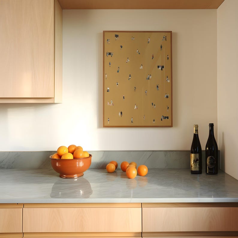 May include: A kitchen scene featuring a terracotta bowl filled with oranges on a gray countertop. A framed artwork with tiny figures hangs on the wall. Two wine bottles stand to the right. Light wood cabinets are visible.