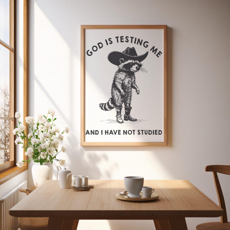 Puede incluir: Un cuadro enmarcado con un mapache con sombrero de vaquero y botas. El texto encima del mapache dice "GOD IS TESTING ME" y debajo "AND I HAVE NOT STUDIED". El cuadro est&aacute; colgado en una pared blanca sobre una mesa de madera.