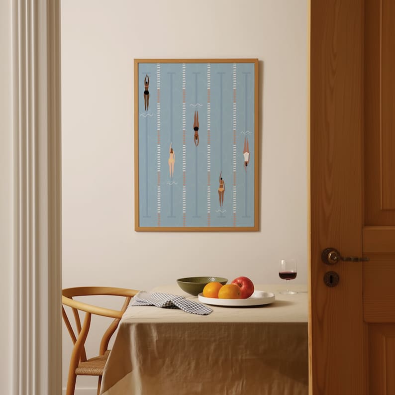 May include: A framed illustration of swimmers in a pool. The swimmers are all women, and they are wearing different colored swimsuits. The pool is blue, and the lanes are marked with white lines. The frame is a light brown wood.