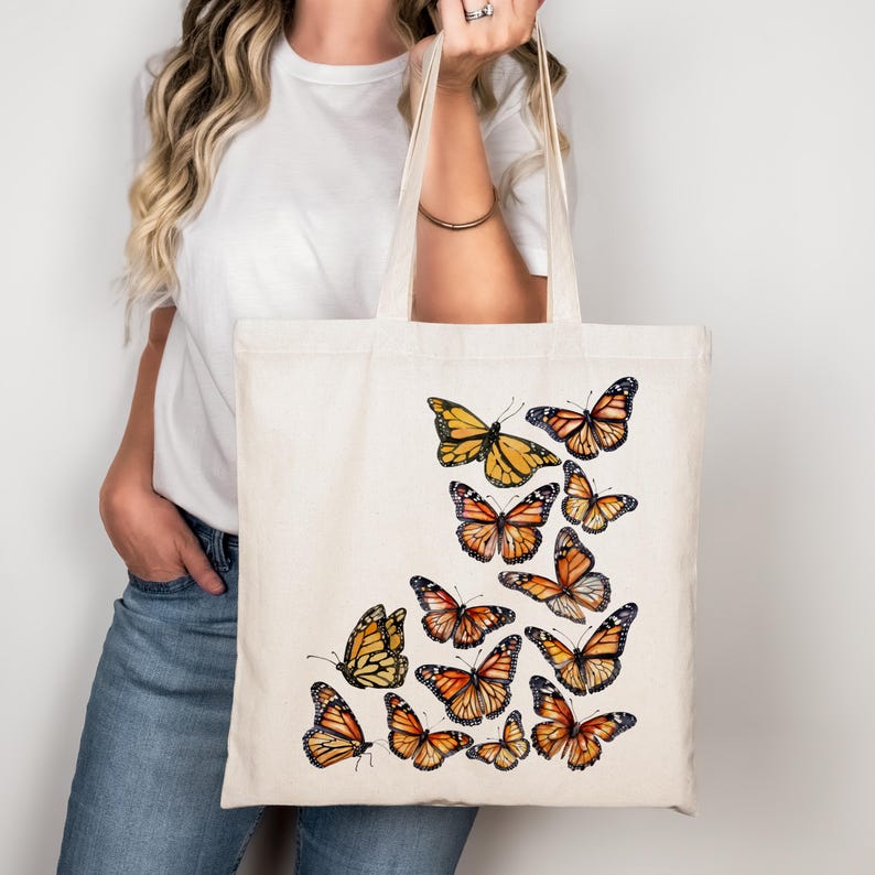 Puede incluir: Un bolso de tela de color crema con un dise&ntilde;o estampado de varias mariposas monarca en tonos naranja, negro y blanco. Las mariposas est&aacute;n dispuestas en un grupo en la parte frontal del bolso, mostrando patrones detallados en sus alas.