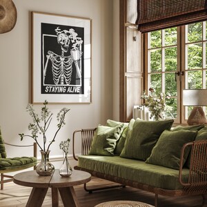 May include: A living room interior with a green velvet sofa, a wooden coffee table, and a framed black and white print of a skeleton drinking coffee with the text "Staying Alive".
