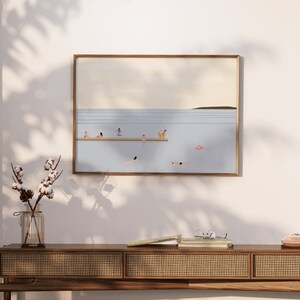 May include: A framed illustration of a beach scene with people swimming and sunbathing. The illustration is in a light blue, beige, and brown color palette. The illustration is hanging on a white wall above a wooden console table with a woven rattan front. A vase with cotton stems is on the table.