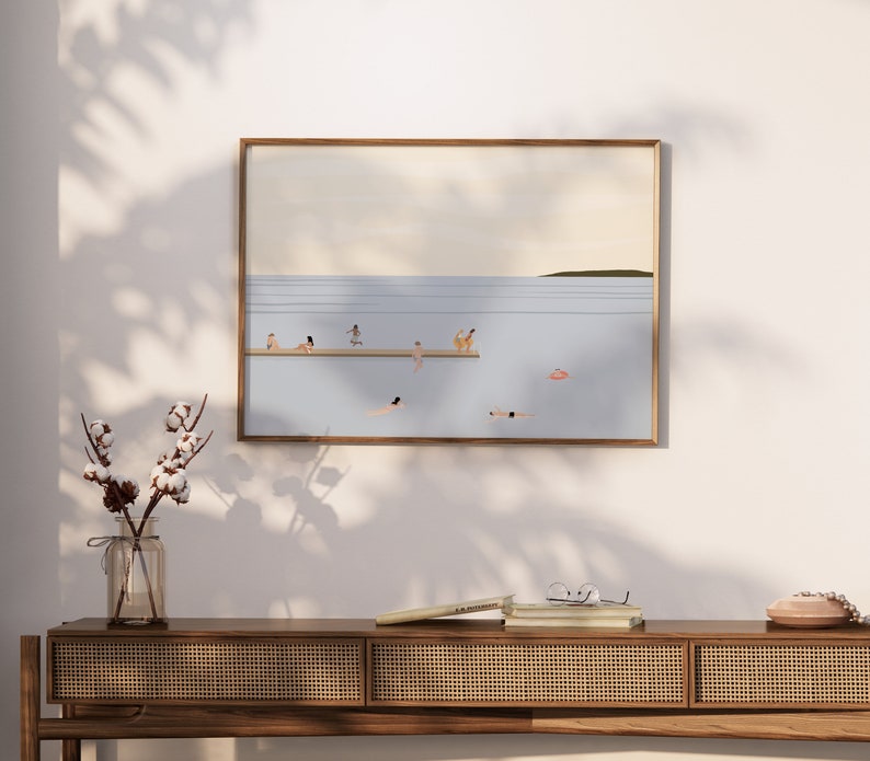 May include: A framed illustration of a beach scene with people swimming and sunbathing. The illustration is in a light blue, beige, and brown color palette. The illustration is hanging on a white wall above a wooden console table with a woven rattan front. A vase with cotton stems is on the table.