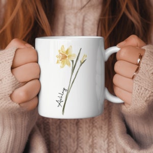May include: White ceramic mug with a watercolor illustration of a yellow daffodil and the name "Ashley" written in cursive.