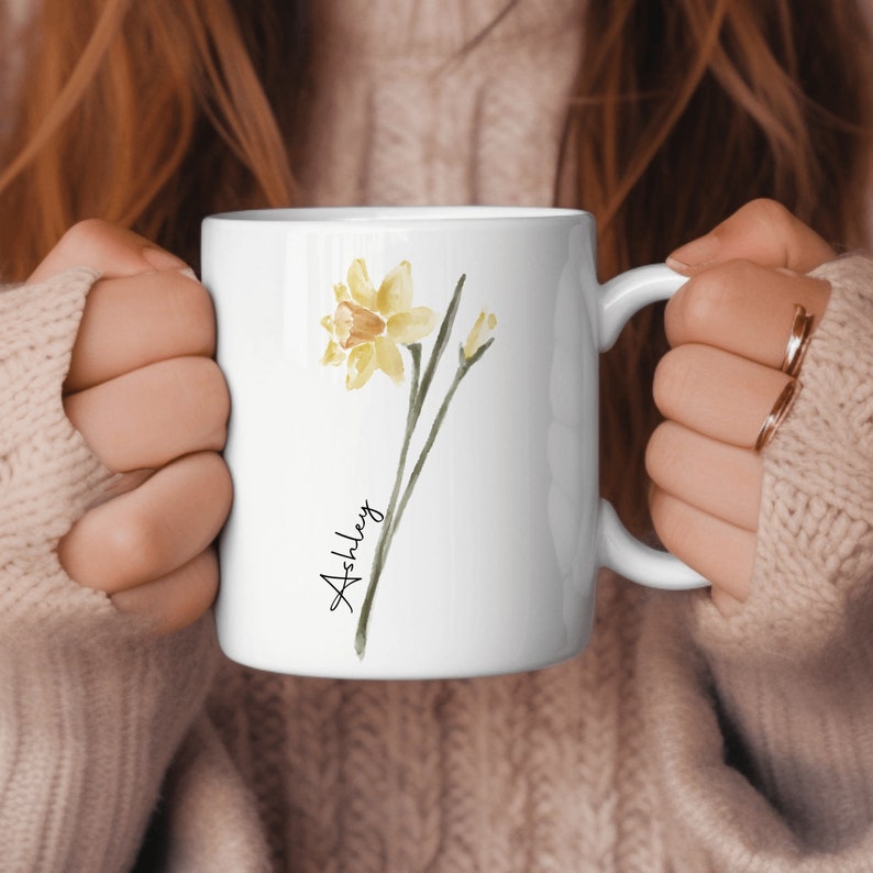 May include: White ceramic mug with a watercolor illustration of a yellow daffodil and the name "Ashley" written in cursive.