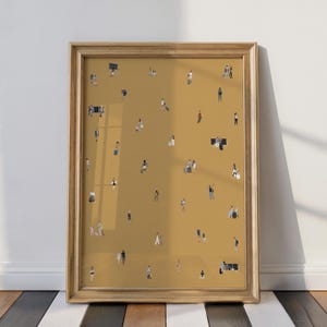 May include: A framed art print featuring a mustard-colored background with numerous tiny figures of people. The figures are dressed in various outfits, some carrying luggage. The frame is a light gold color, and the artwork is displayed against a white wall.