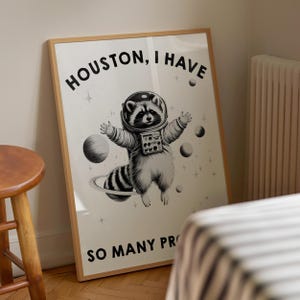 May include: A framed wall art print featuring a black and white illustration of a raccoon wearing an astronaut helmet and suit, floating in space among planets and stars. The text reads HOUSTON, I HAVE SO MANY PROB at the top and bottom.