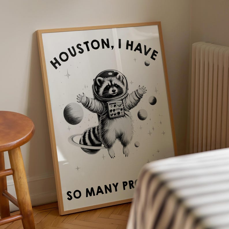 May include: A framed wall art print featuring a black and white illustration of a raccoon wearing an astronaut helmet and suit, floating in space among planets and stars. The text reads HOUSTON, I HAVE SO MANY PROB at the top and bottom.