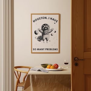 May include: A rectangular wall art print in a light wood frame featuring a black and white illustration of a raccoon wearing a space suit and helmet floating in space with planets and stars, with the text HOUSTON, I HAVE SO MANY PROBLEMS.