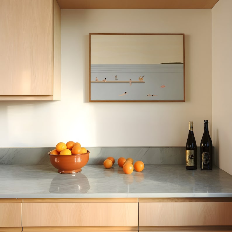 May include: A kitchen counter with a brown bowl filled with orange fruit, two bottles of wine, and a framed picture of a beach scene with people swimming and sunbathing.