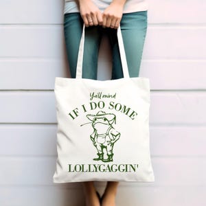 May include: White canvas tote bag with green text and a cartoon frog wearing a hat. The text reads "Y'all mind if I do some lollygaggin'"