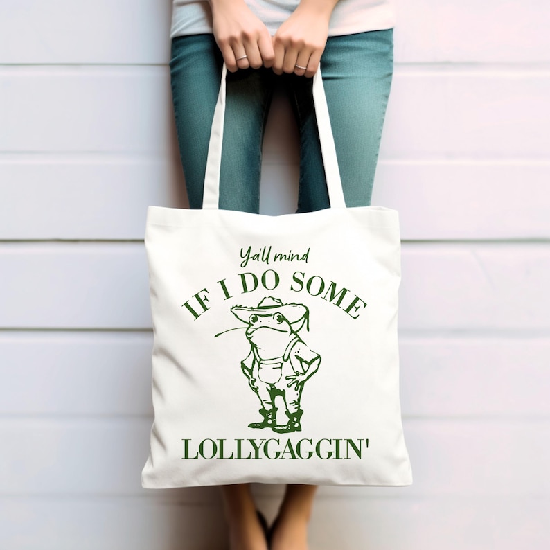May include: White canvas tote bag with green text and a cartoon frog wearing a hat. The text reads "Y'all mind if I do some lollygaggin'"