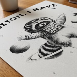 May include: A black and white art print featuring a raccoon wearing a space suit and helmet, floating in space near a planet with rings. The text "HOUSTON, I HAVE" is printed in black at the top. The design includes stars and small planets.