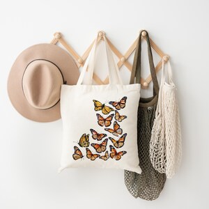 Puede incluir: Un bolso de tela de color crema con un dise&ntilde;o estampado de varias mariposas monarca de color naranja y negro. Las mariposas est&aacute;n dispuestas en un grupo en la parte frontal del bolso, mostrando varios patrones de alas y tama&ntilde;os en colores vivos.