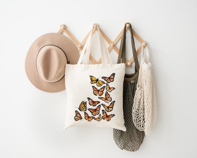 Puede incluir: Un bolso de tela de color crema con un dise&ntilde;o estampado de varias mariposas monarca de color naranja y negro. Las mariposas est&aacute;n dispuestas en un grupo en la parte frontal del bolso, mostrando varios patrones de alas y tama&ntilde;os en colores vivos.