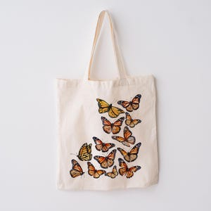 Puede incluir: Una bolsa de tela de color crema con un dise&ntilde;o estampado de varias mariposas monarca en tonos naranja, negro y blanco. Las mariposas est&aacute;n dispuestas en un grupo en la parte inferior derecha de la bolsa, que cuenta con dos asas.