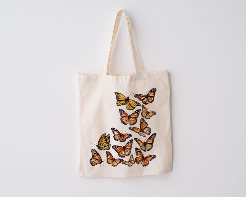 Puede incluir: Una bolsa de tela de color crema con un dise&ntilde;o estampado de varias mariposas monarca en tonos naranja, negro y blanco. Las mariposas est&aacute;n dispuestas en un grupo en la parte inferior derecha de la bolsa, que cuenta con dos asas.