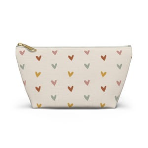 May include: A cream-colored zippered pouch featuring a repeating pattern of small, hand-drawn style hearts in muted shades of terracotta, sage green, dusty pink, and mustard yellow, finished with a gold-toned metal zipper pull at the top.