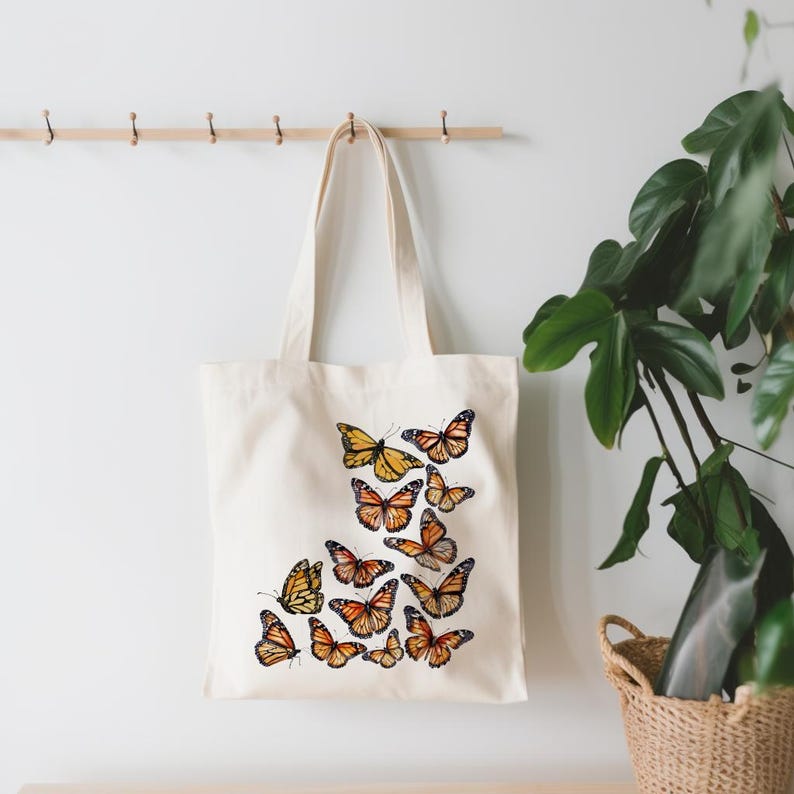 Puede incluir: Un bolso de tela de color crema con un dise&ntilde;o estampado de varias mariposas monarca en tonos naranja, negro y blanco. Las mariposas est&aacute;n dispuestas en un grupo en la parte inferior derecha del bolso, el cual cuenta con dos asas.