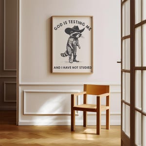 Puede incluir: Un cuadro enmarcado con un mapache con sombrero y botas de vaquero. El texto dice: "GOD IS TESTING ME AND I HAVE NOT STUDIED". El cuadro est&aacute; colgado sobre una silla de madera.